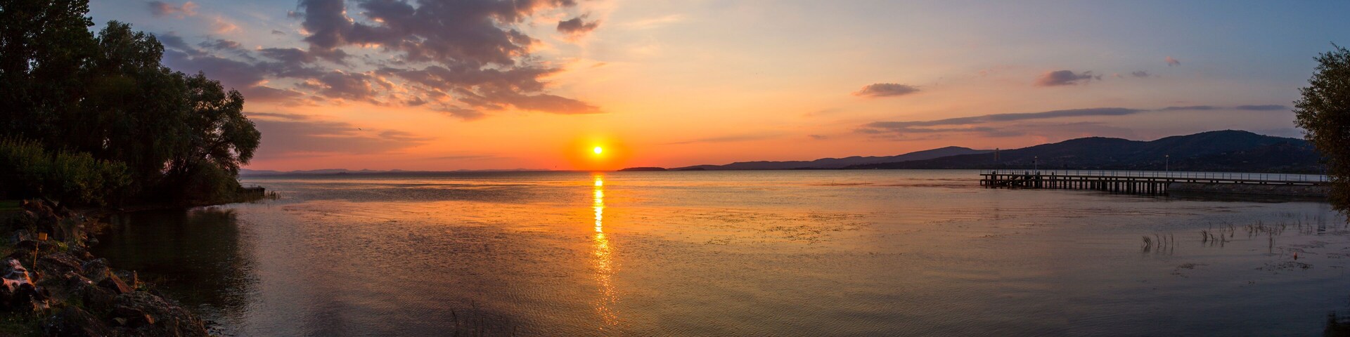 Um pôr do sol sobre um lago tranquilo, com um cais que se estende na água, rodeado por árvores e colinas ao longe.