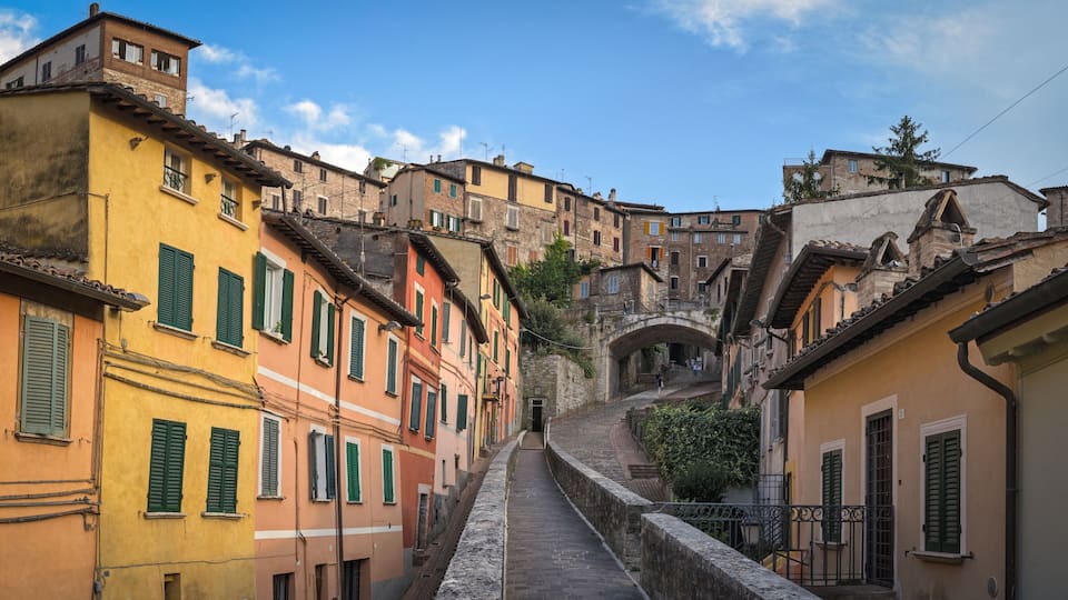 A narrow street flanked by colorful buildings with green shutters, leading up to a stone bridge.