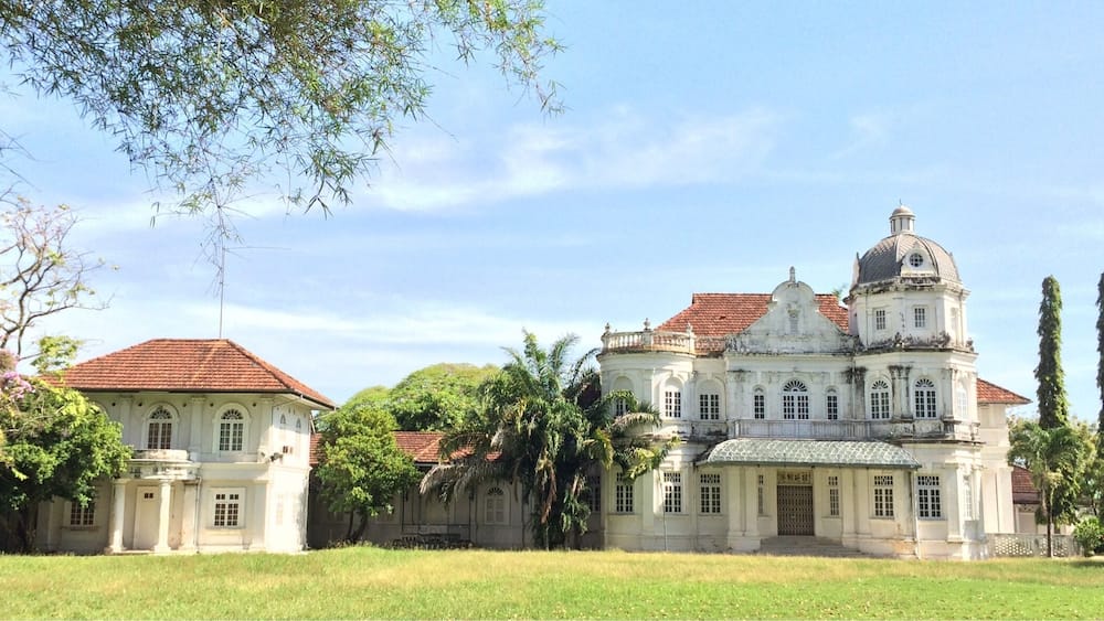 A large colonial-style building with a red-tiled roof, surrounded by lush greenery and palm trees.