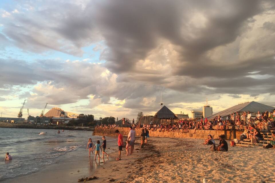A crowded beach with people walking and sitting, a large crowd gathered near a stage, and a partly cloudy sky.