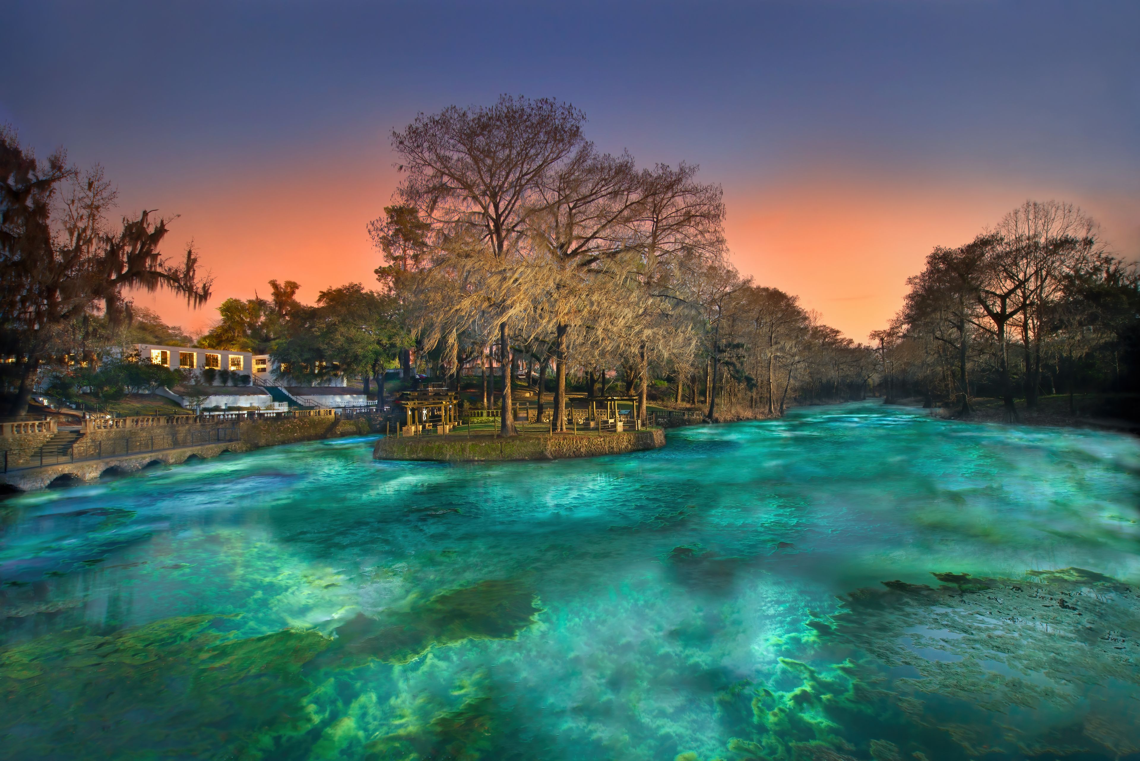 A river with clear turquoise water, a stone bridge, and a gazebo surrounded by trees.