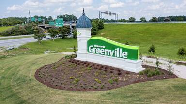 Un letrero de bienvenida a Greenville con una paleta de colores verde y blanco, rodeado de un área ajardinada con árboles y arbustos pequeños.