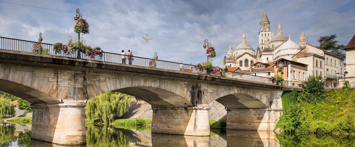 A stone bridge over a calm river with a reflection, a church with domes, and hanging flower baskets.