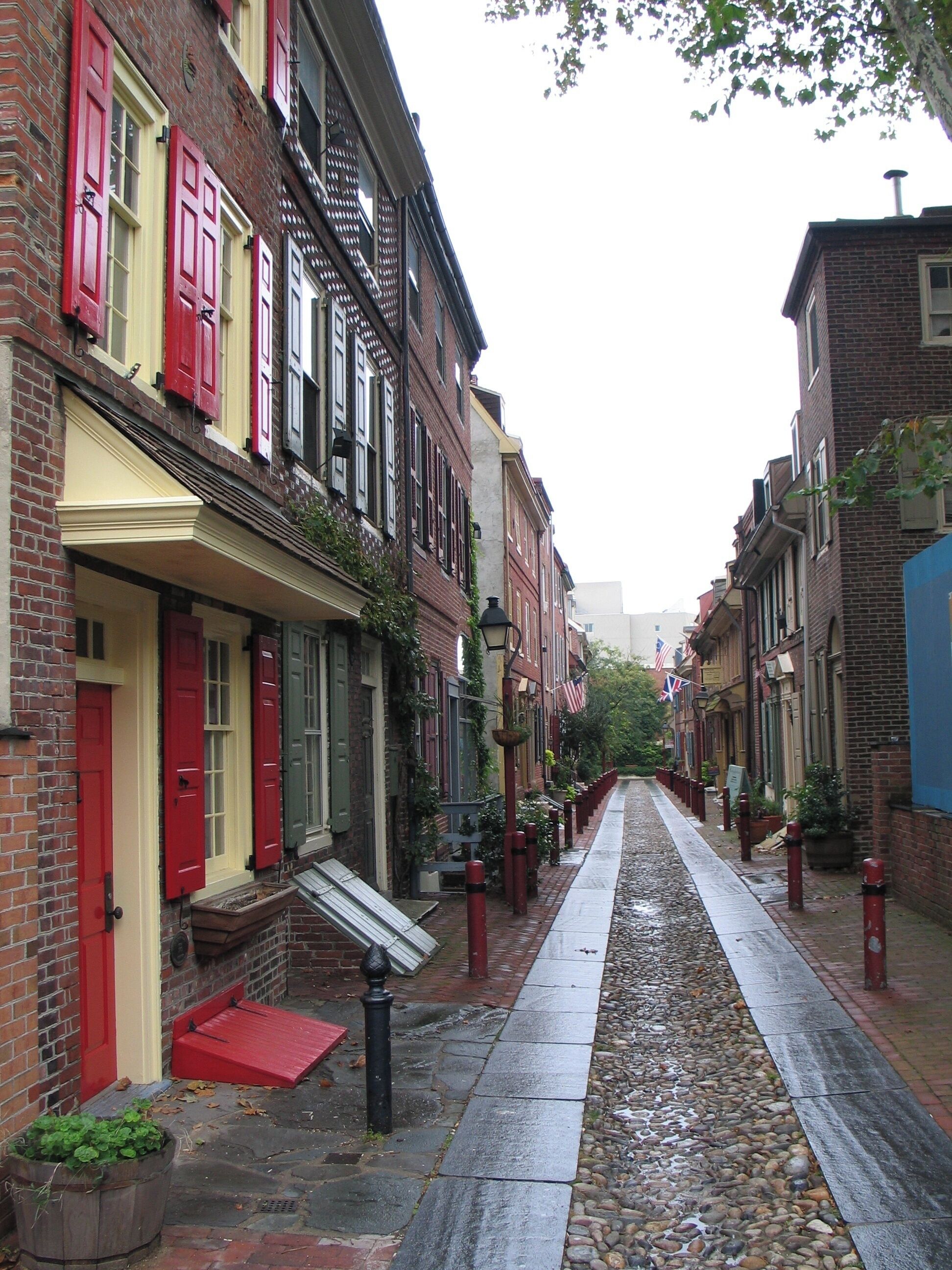 Una estrecha calle empedrada flanqueada por edificios de ladrillo, contraventanas rojas y banderas estadounidenses.