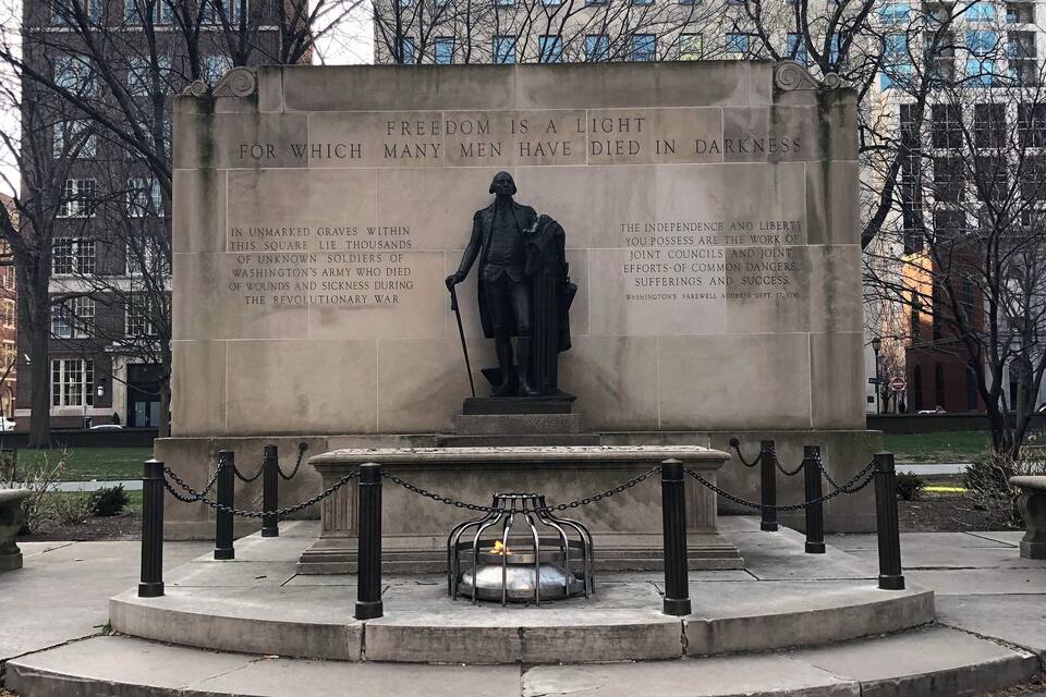 Um memorial com estátua e inscrições, rodeado por um pavimento e grades de corrente.