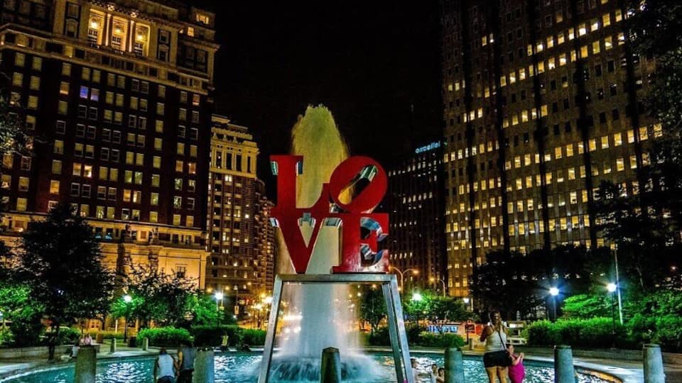 A nighttime cityscape with a large illuminated sculpture of the letters 'LOVE' in the foreground, surrounded by a fountain and modern buildings.