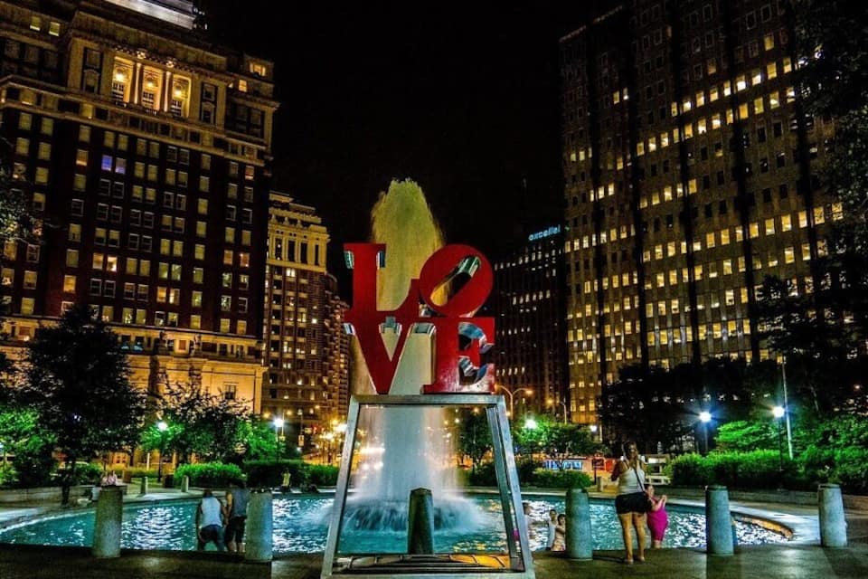 Une vue nocturne d’une ville, avec une grande sculpture illuminée représentant les lettres « LOVE » au premier plan, entourée d’une fontaine et de bâtiments modernes.