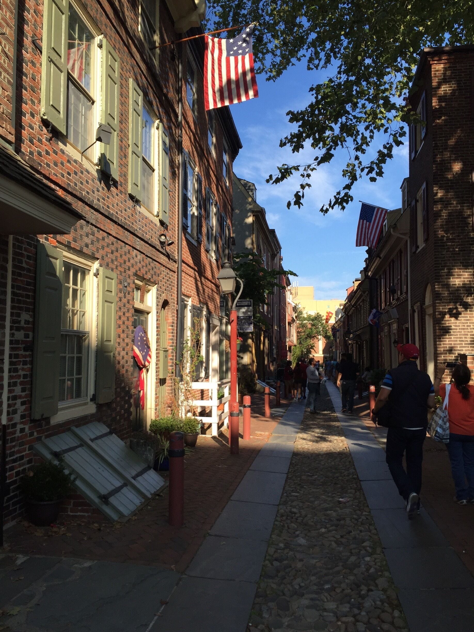 A cobblestone street lined with brick buildings, American flags, and pedestrians.