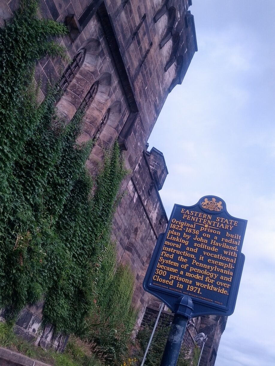 Placa histórica fixada numa parede de pedra coberta de hera, detalhando a história da Penitenciária Estadual do Leste.