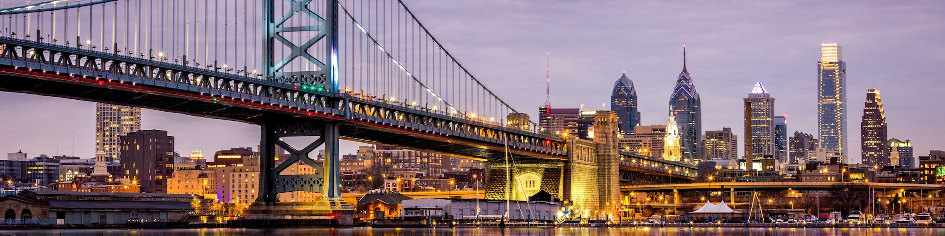 Un puente colgante sobre un río tranquilo, con un horizonte urbano al fondo.
