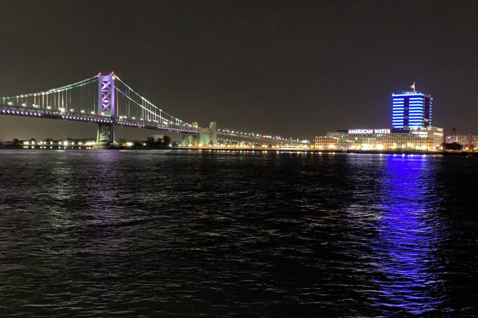 A bridge with illuminated lights reflecting on the water, a building with 'AMERICAN WATER' sign, and a city skyline in the background.