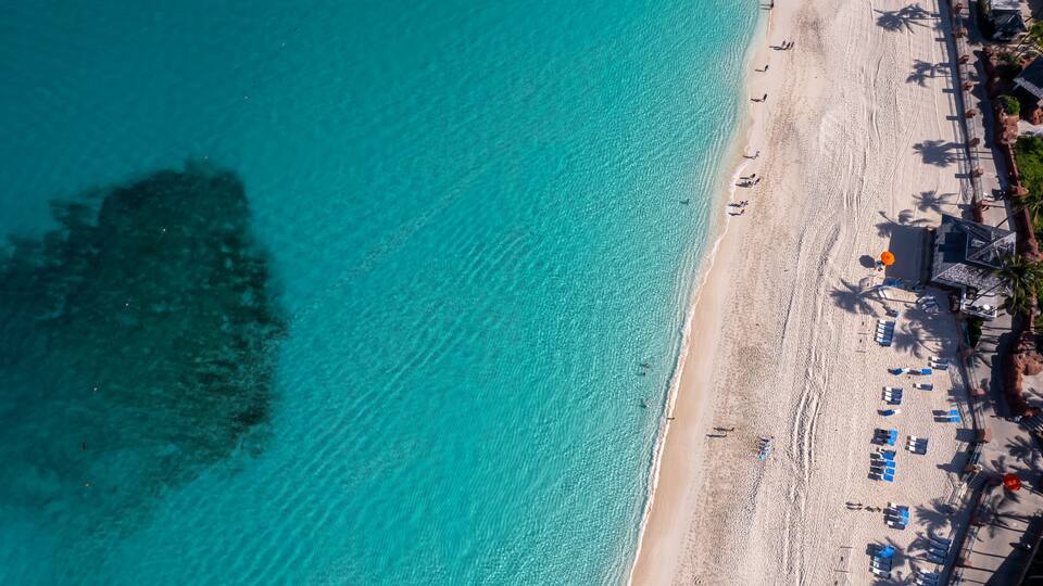 A turquoise beach with clear water, a sandy shore, and a row of beach umbrellas and lounge chairs.