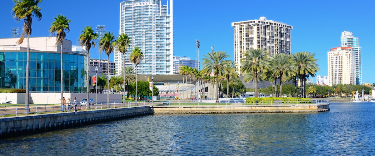 A waterfront cityscape with modern high-rise buildings, palm trees, and a bridge over the water.