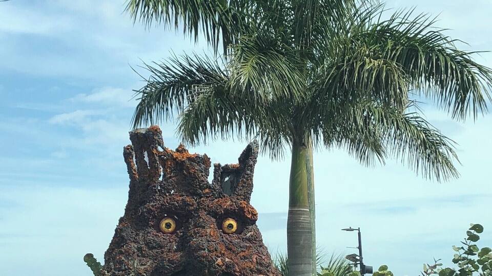 A large, rust-colored sculpture resembling a creature with large eyes and a textured surface, surrounded by greenery and a palm tree.