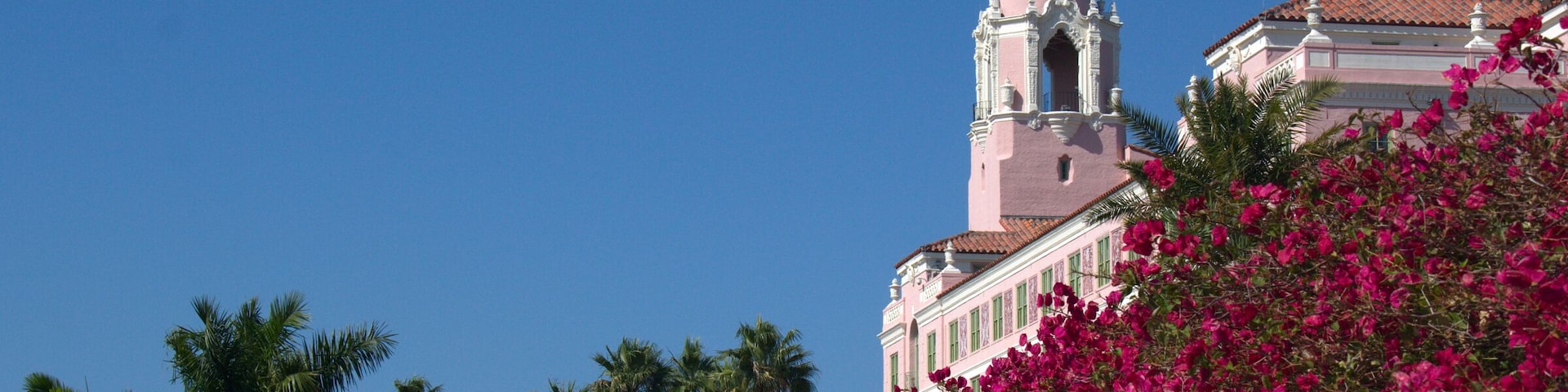 Un edificio rosa con una torre de campanas, rodeado de buganvillas rosadas y palmeras.
