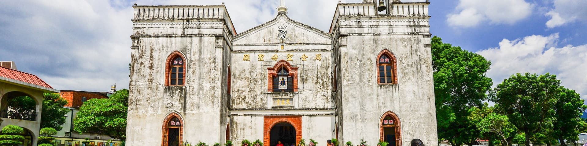 A historic church with a large central archway and two towers, surrounded by a paved courtyard and greenery.