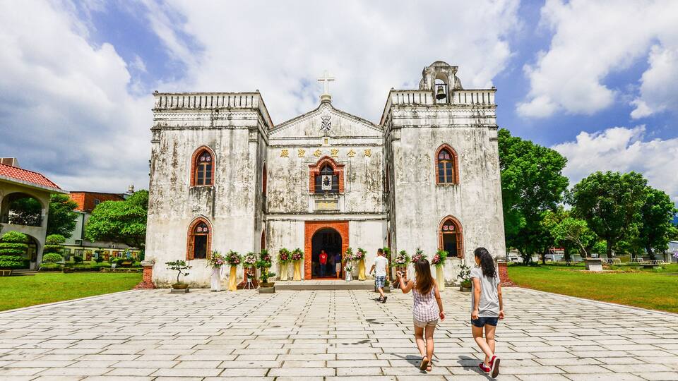 A historic church with a large central archway and two towers, surrounded by a paved courtyard and greenery.