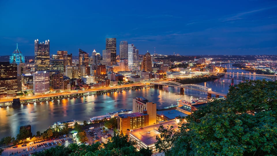 A city skyline at dusk with illuminated buildings, a river, and a bridge.