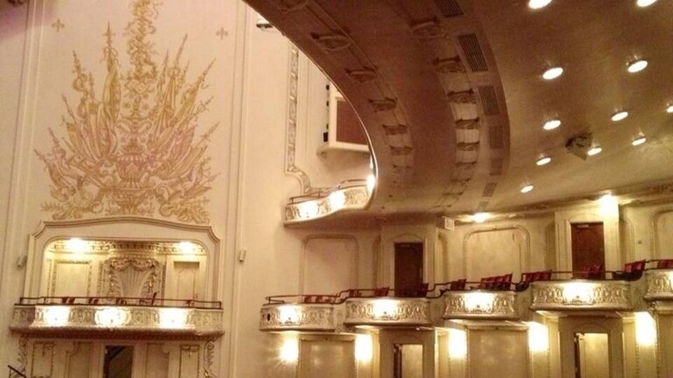 An ornate theater interior with rows of red seats, a large chandelier, and a decorative wall mural.
