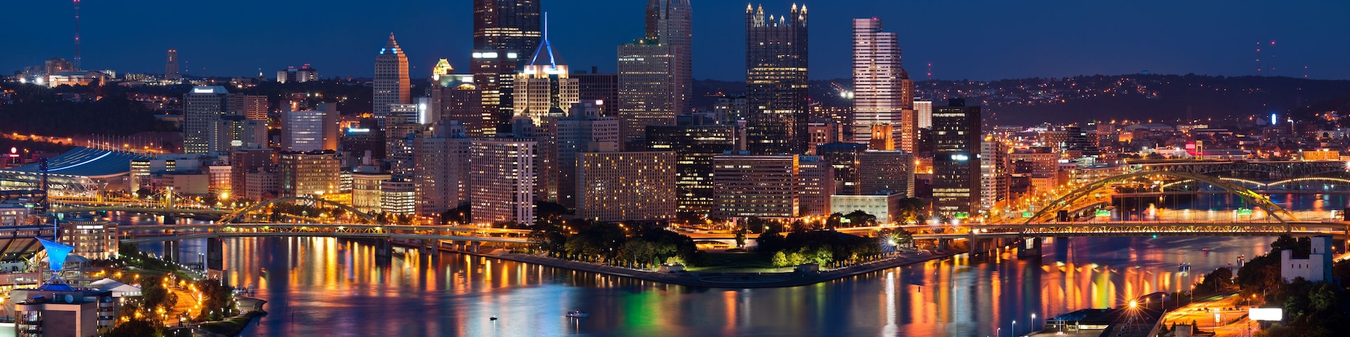 A city skyline at night with illuminated buildings and a river reflecting the lights.