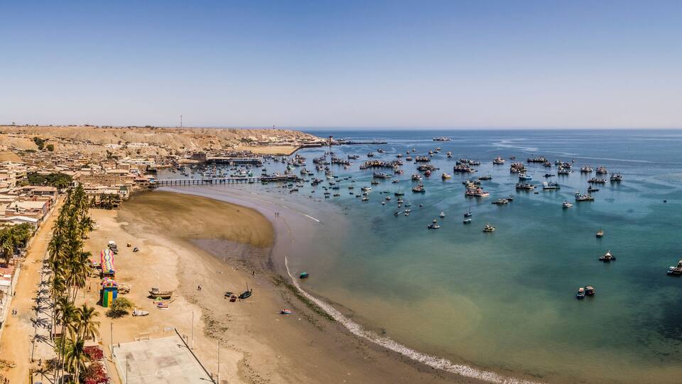 A coastal town with a sandy beach, a marina with numerous boats, and a hilly landscape in the background.