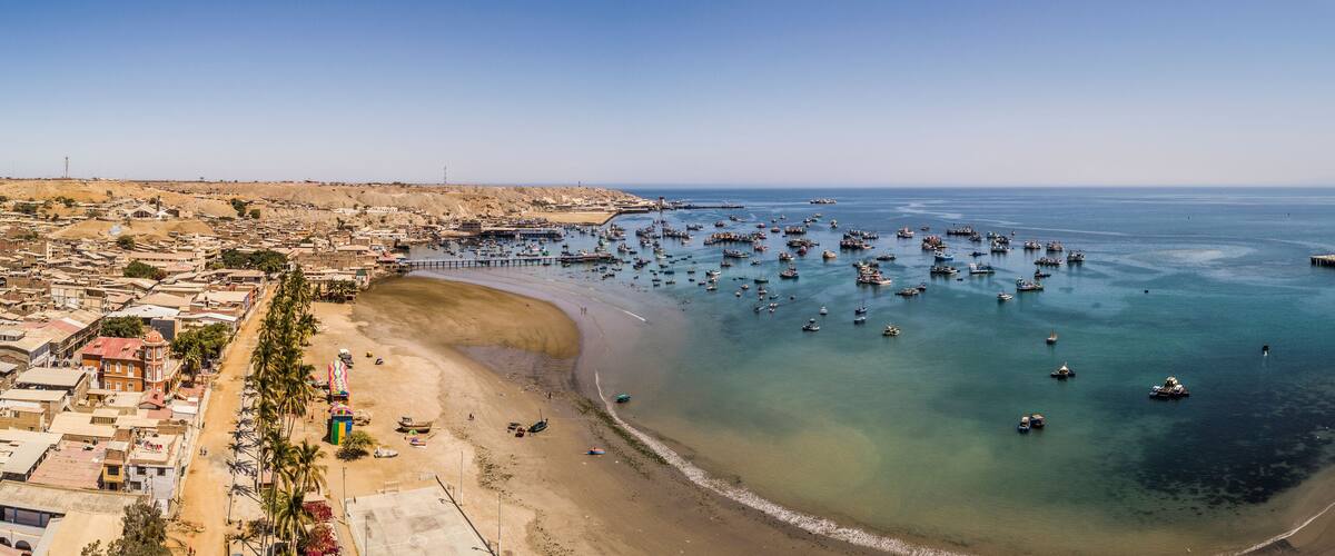 A coastal town with a sandy beach, a marina with numerous boats, and a hilly landscape in the background.