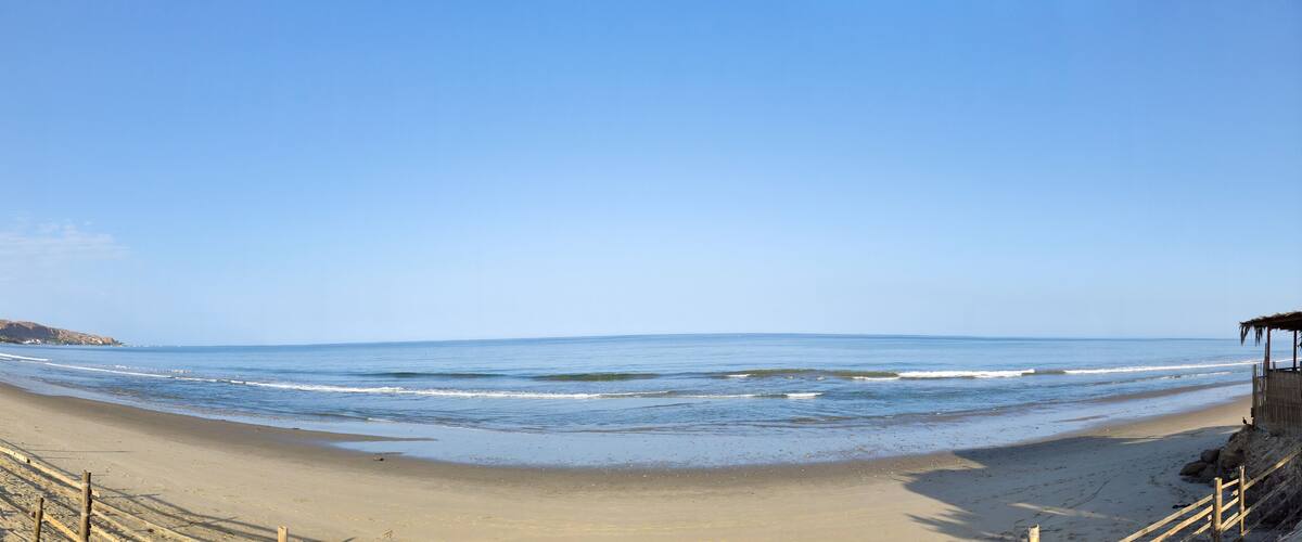 A beach with a wooden fence, a small hut, and a rocky outcrop in the distance.