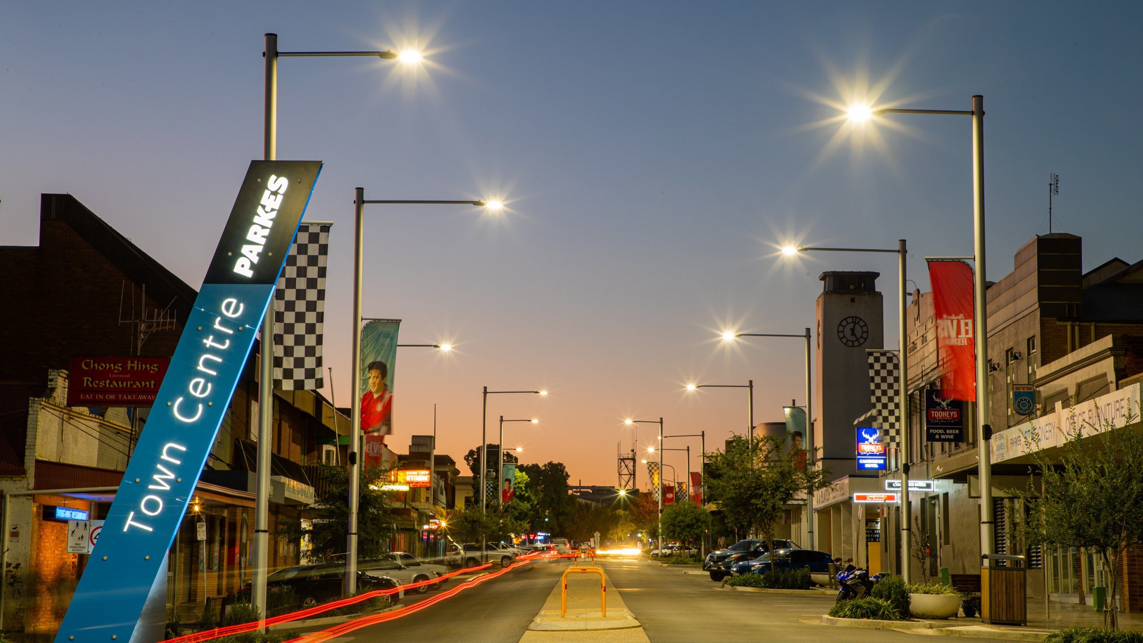 Rua urbana ao entardecer, com luminárias iluminadas, uma faixa azul com a inscrição 'Town Centre' e diversas lojas.