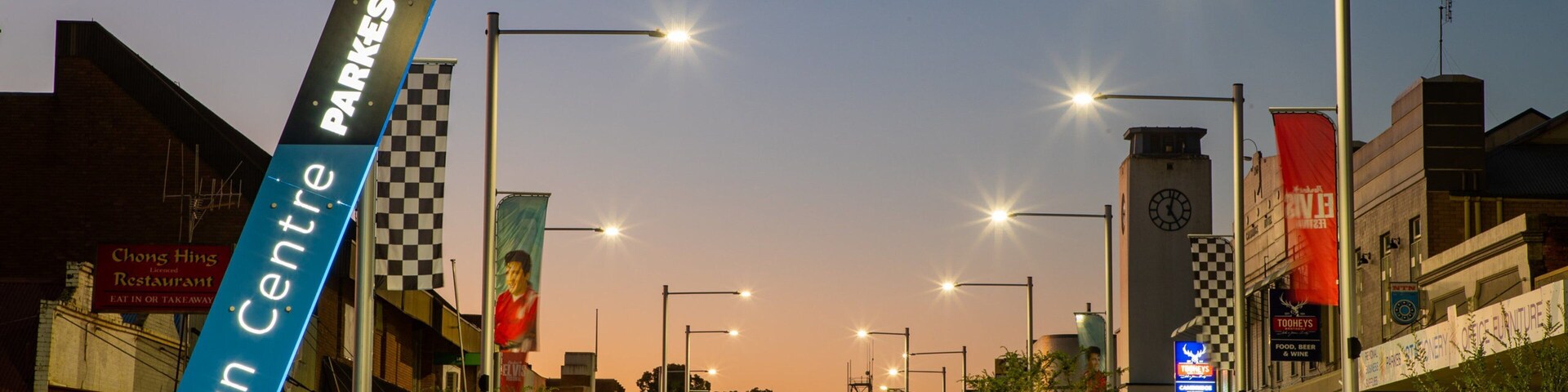 Rua urbana ao entardecer, com luminárias iluminadas, uma faixa azul com a inscrição 'Town Centre' e diversas lojas.