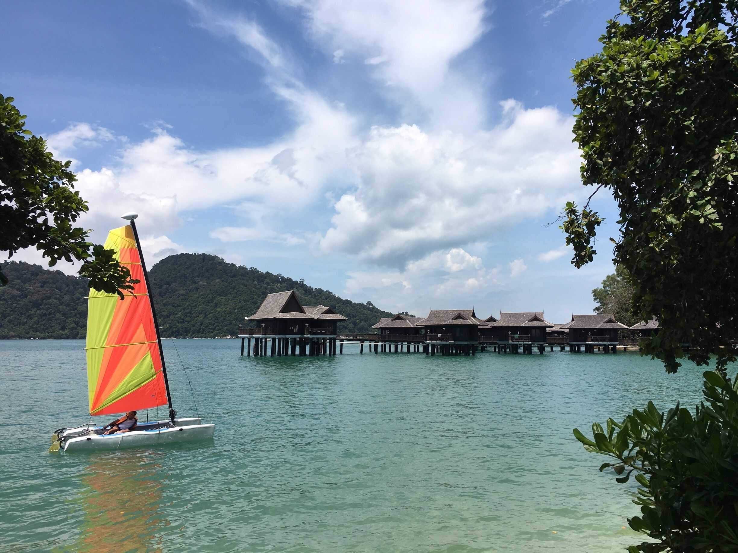 Un velero con vela de colores en aguas turquesa, rodeado de bungalows sobre el agua y exuberante vegetación.