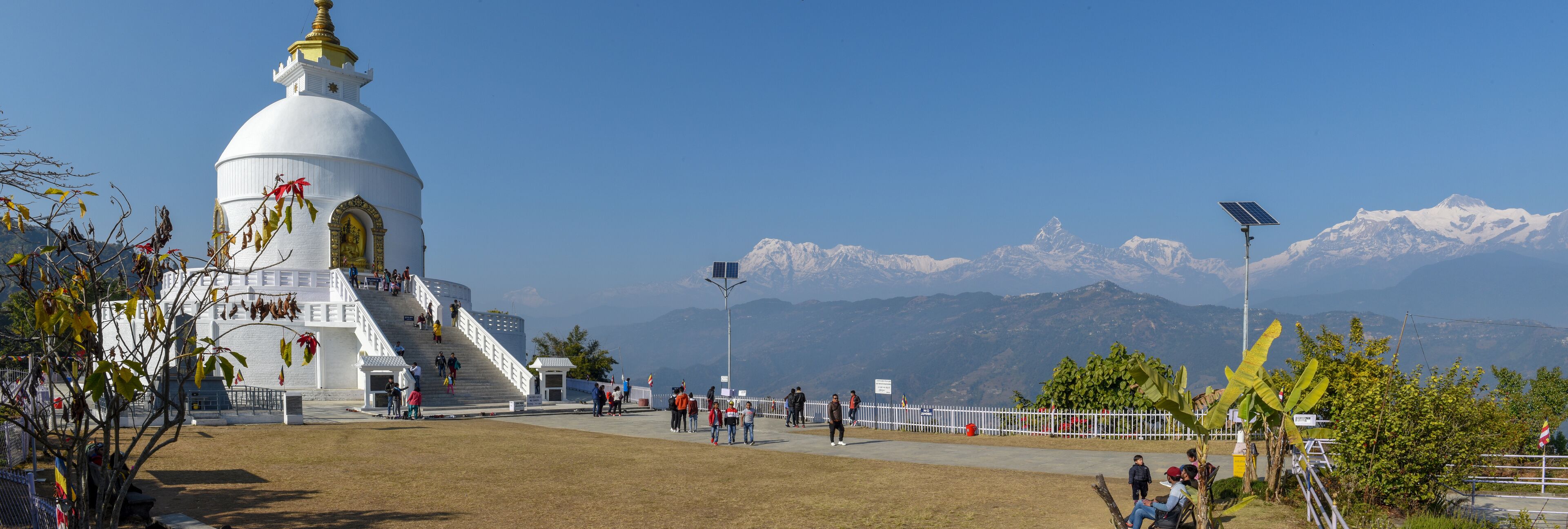 A white temple with a golden spire, surrounded by a fenced area with people walking and a mountain range in the background.