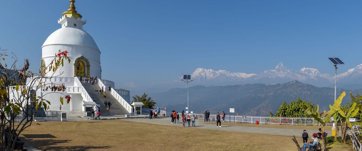 A white temple with a golden spire, surrounded by a fenced area with people walking and a mountain range in the background.