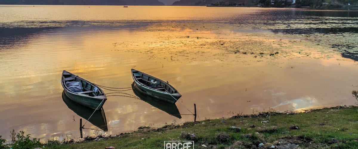 Two small boats float peacefully close to the shore of Phewa Lake, right before the Sun disappears behind the mountains that surround it.