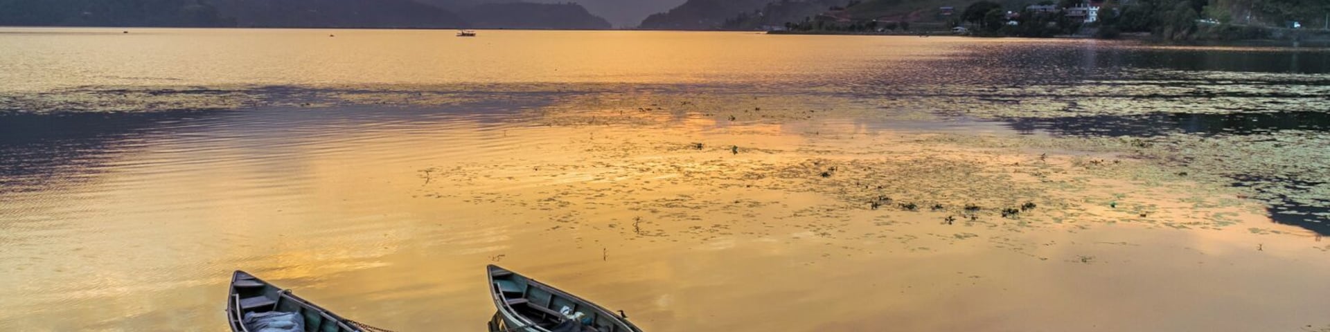 Two small boats float peacefully close to the shore of Phewa Lake, right before the Sun disappears behind the mountains that surround it.