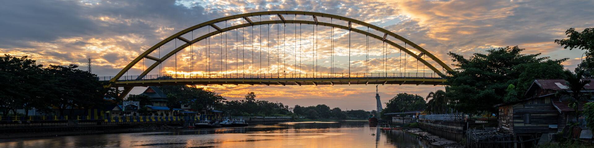 A bridge over a calm river reflecting the sky, surrounded by trees and buildings.