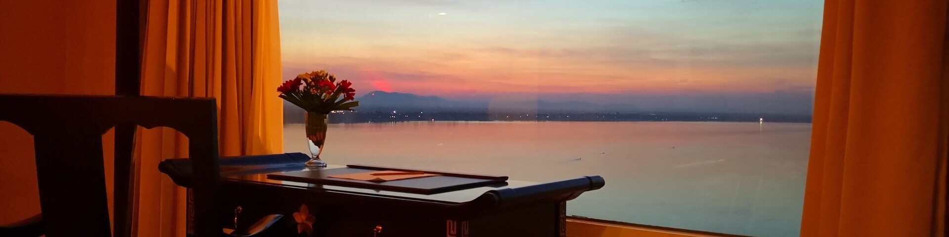 Uma mesa com um vaso de flores e um menu, uma cadeira e vista para um corpo d'água calmo sob um céu com o pôr do sol.