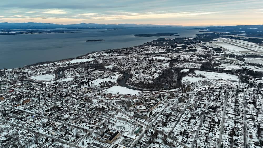 Une vue aérienne d’une ville enneigée, avec une rivière, des ponts et des montagnes à l’horizon.