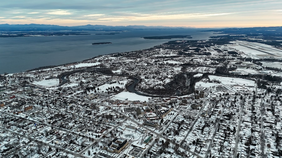 An aerial view of a snow-covered city with a river, bridges, and distant mountains.