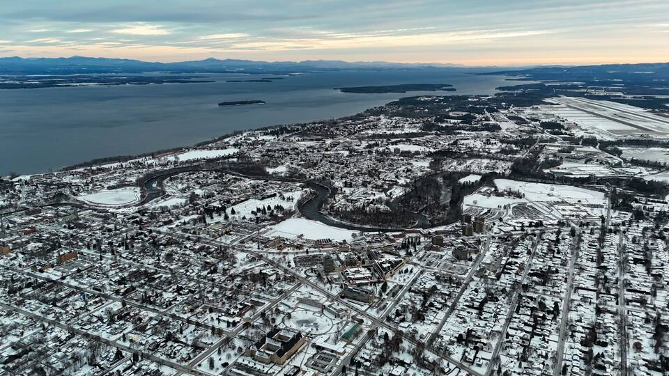 An aerial view of a snow-covered city with a river, bridges, and distant mountains.