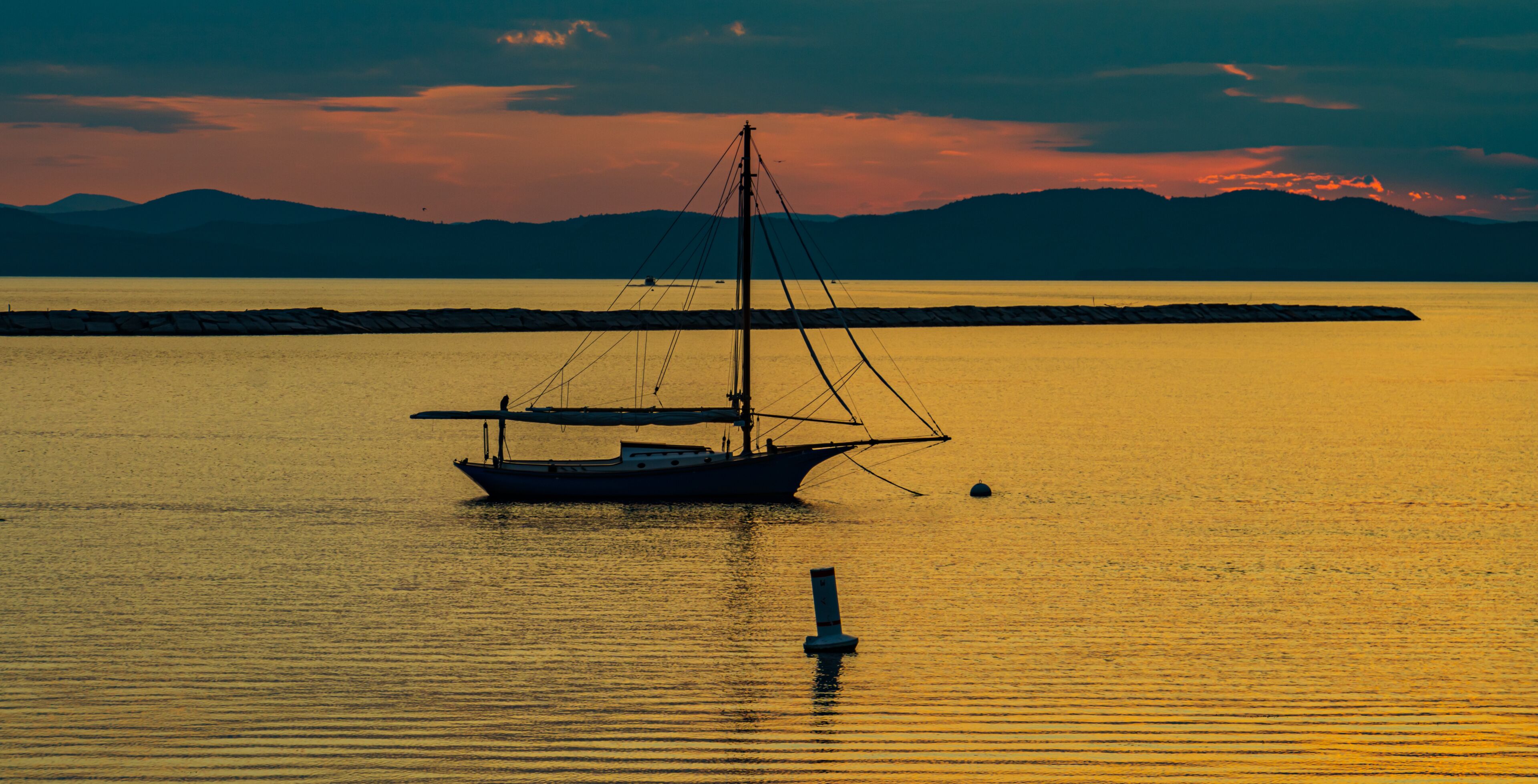 banner photo of sloop sail boat moored  on the shores of Lake Champlain in Vermont as the sunsets behind the Adirondack Mountains in New York across the lake 
