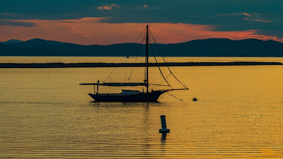 banner photo of sloop sail boat moored on the shores of Lake Champlain in Vermont as the sunsets behind the Adirondack Mountains in New York across the lake