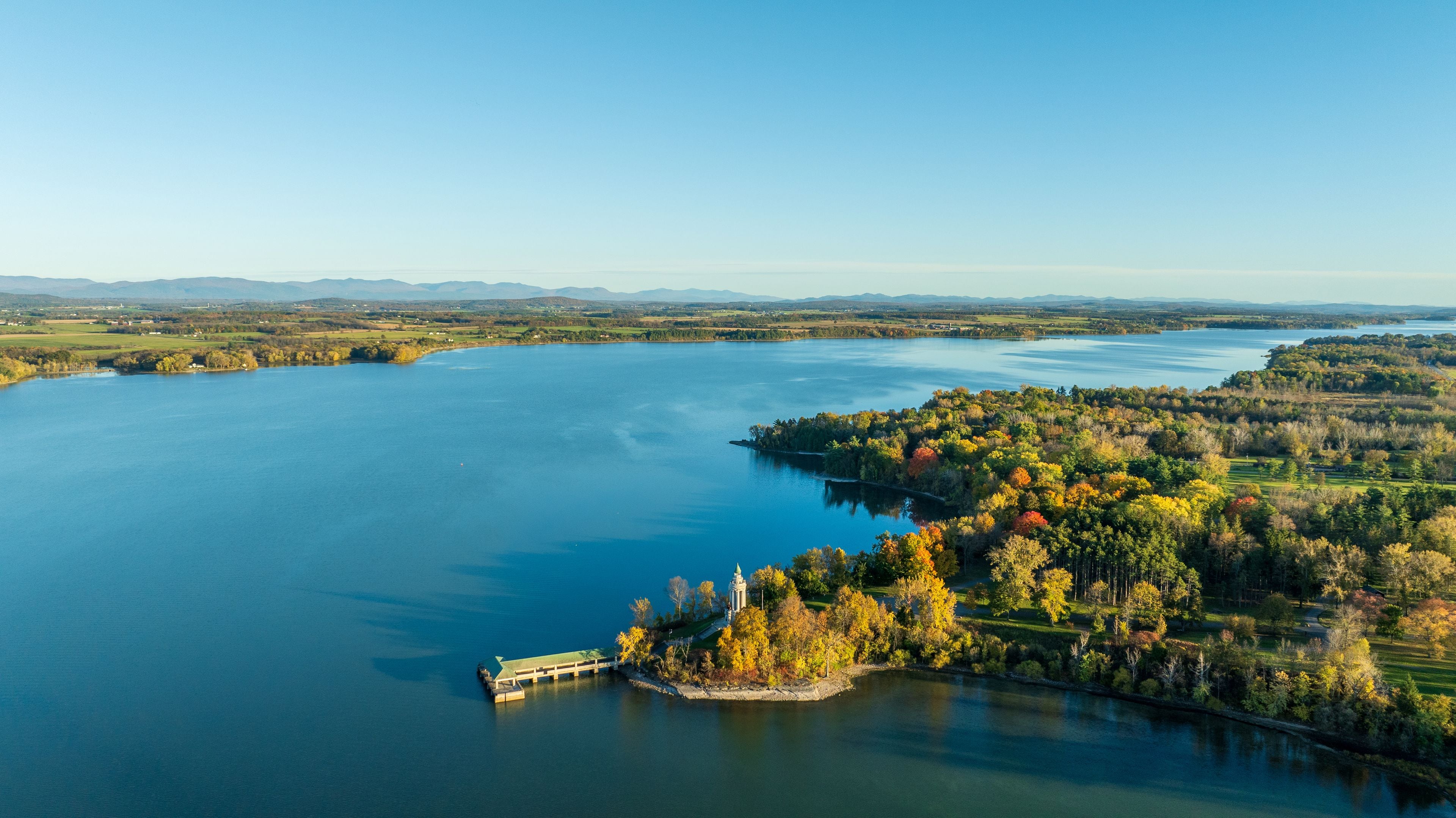 A large body of water with a dock, surrounded by autumn-colored trees and distant mountains.