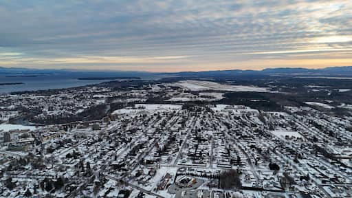 Plattsburgh cityscape receiving fresh snowfall, showcasing residential neighborhoods with snow covered roofs and streets, extending towards lake champlain and distant mountains under a dramatic sky
