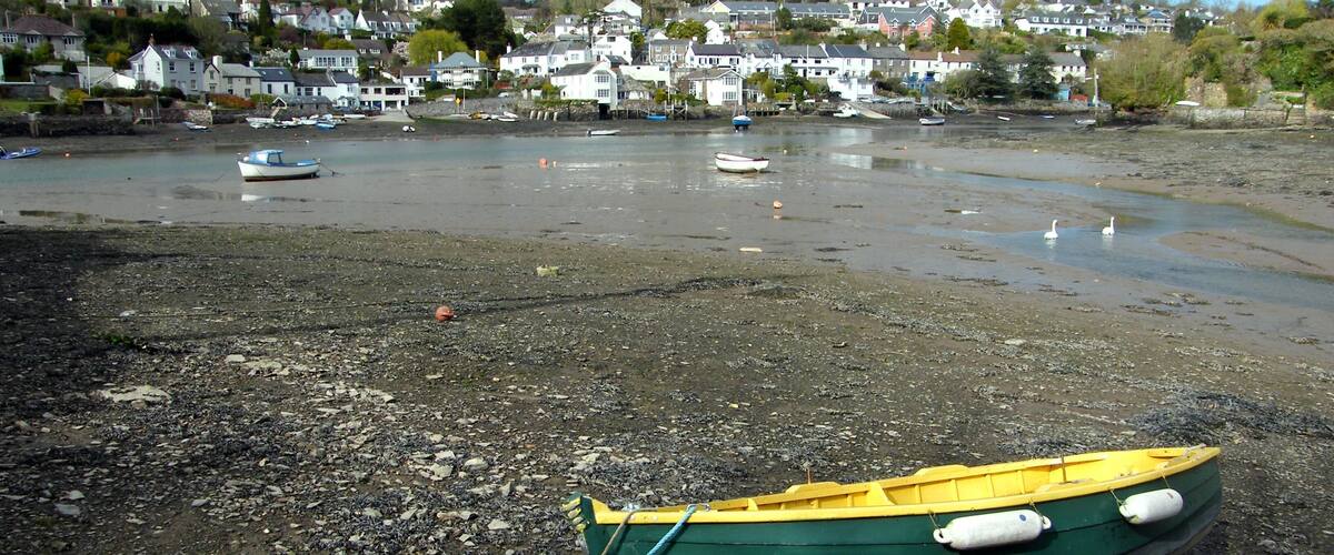 A rocky beach with a green and yellow boat tied to a buoy, surrounded by calm water and houses in the background.