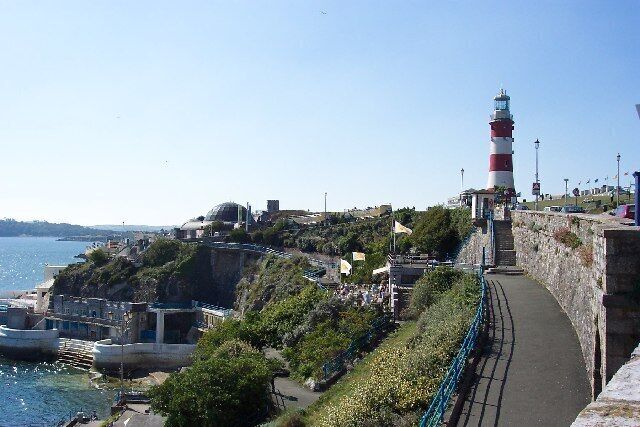 Un paseo marítimo con un faro, un muro de piedra y barcos amarrados en el muelle.