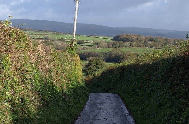 A narrow rural road flanked by lush greenery and rolling hills in the distance.