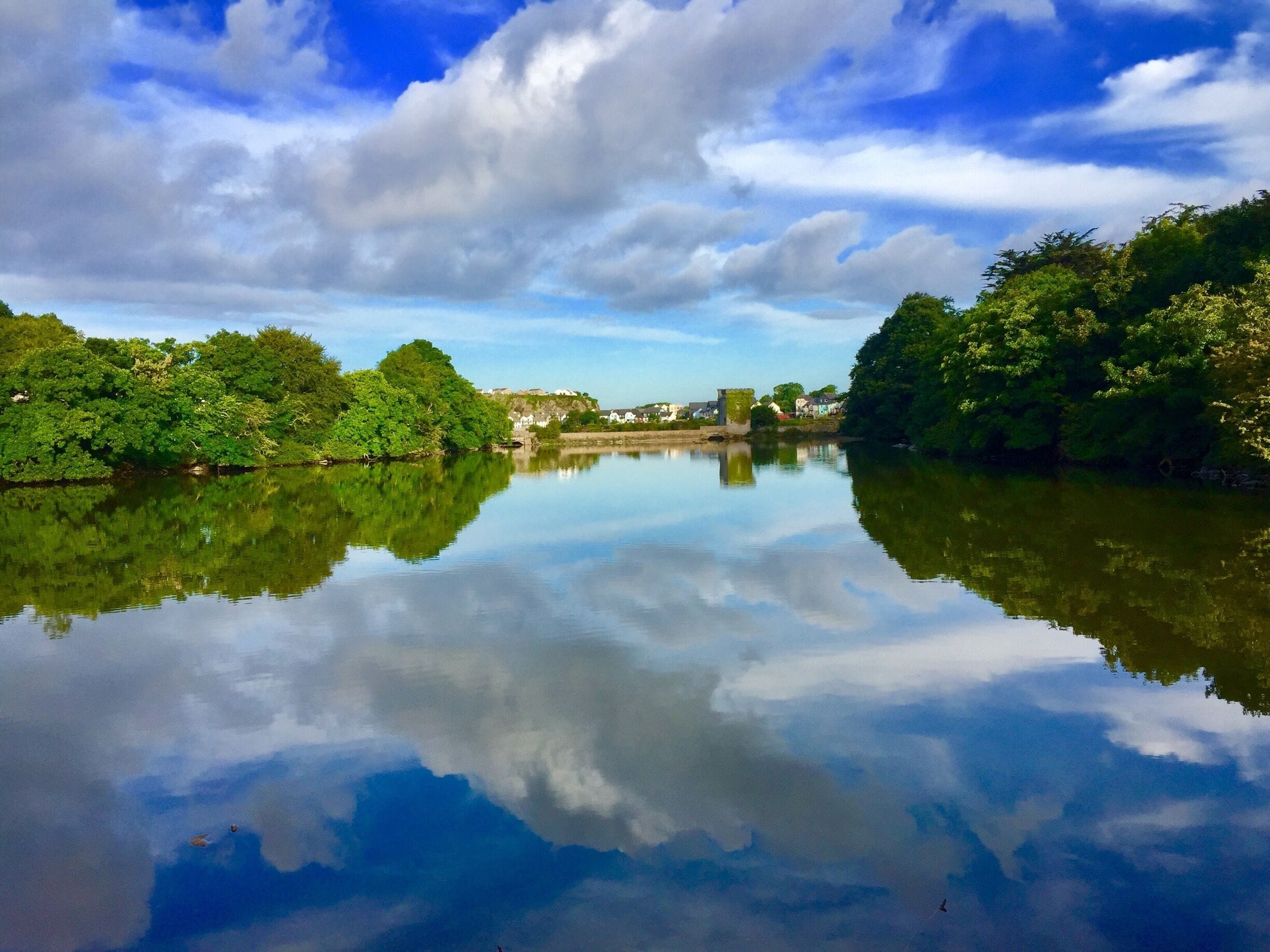 A calm river reflecting trees and a distant building.