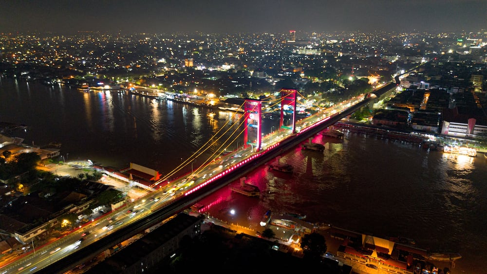 A brightly lit bridge with red supports over a river, surrounded by a cityscape at night.