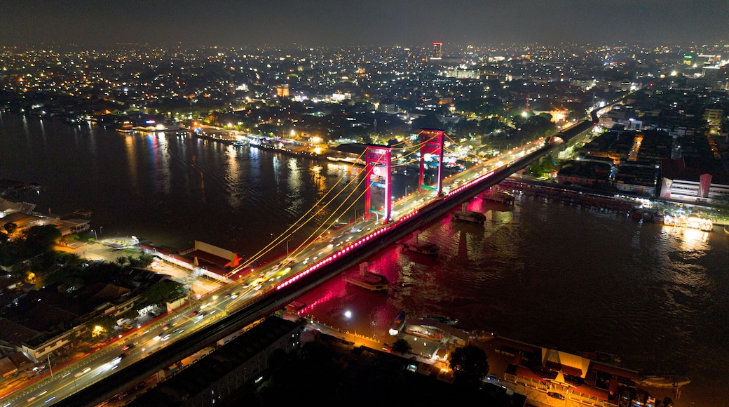 Un pont illuminé de mille feux, avec des piliers rouges, enjambant une rivière, et entouré d’un paysage urbain nocturne.