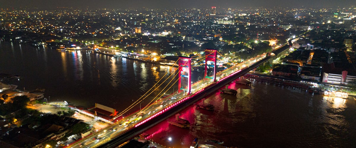 A brightly lit bridge with red supports over a river, surrounded by a cityscape at night.